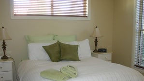 A neatly made bed with a white comforter and two green pillows, a white nightstand with a lamp, and a window with blinds.