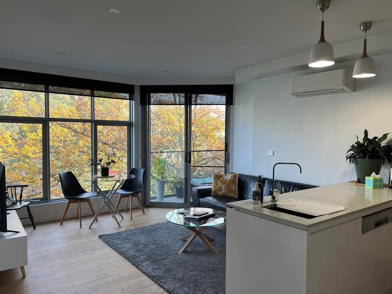 A modern kitchen with a glass island, a dining area with chairs, a sofa, and a large window with autumn foliage view.