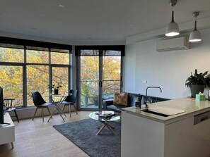 Et moderne køkken med en glasø, et spiseområde med stole, en sofa og et stort vindue med udsigt til efterårsfarver.