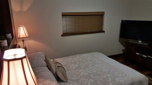 Une chambre à coucher avec un lit, des oreillers, une lampe, une fenêtre avec des stores et un téléviseur posé sur un meuble.