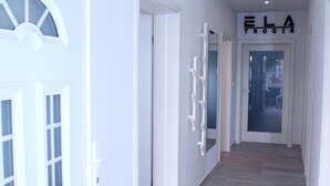 A hallway with a wooden floor, white walls, and a door with a frosted glass window. There is a sign above the door that reads "ELA TROGIR".