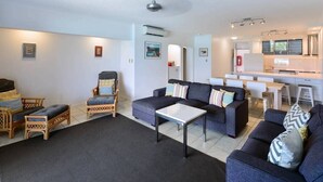 A modern living room with a sofa, chairs, a coffee table, and a kitchen area with a counter and appliances. - Frangipani Lodge 008, Beachfront Apartment on Hamilton Island (Hamilton Island)