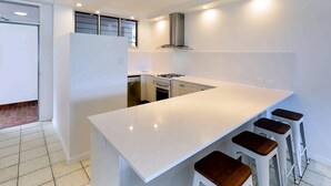 A modern kitchen with a white countertop, a gas stove, and a stainless steel range hood. There are three stools at the counter. - Frangipani Lodge 008, Beachfront Apartment on Hamilton Island (Hamilton Island)