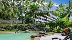 A pool area with lounge chairs, palm trees, and a building in the background. - Frangipani Lodge 008, Beachfront Apartment on Hamilton Island (Hamilton Island)