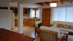 Una sala de estar acogedora con sofá de madera y una mesa de comedor, una cocina con armarios de madera, y un ventanal con cortinas.