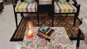 A living room with a sofa set, a table with books and a candle, and a ceiling fan. - Colonels Home stay - A home away from home. Set in a scenic natural environment. (Sector 14)