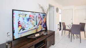 A modern living room with a flat-screen TV, dark wooden entertainment unit, and a dining area with a small table and chairs.