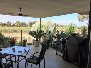 Un patio techado con mesa y sillas, ventilador de techo y vista a un jardín ajardinado y casas al fondo.