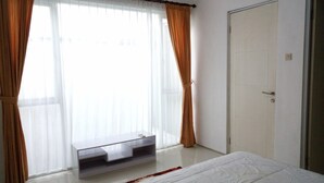 Un dormitorio con cama, ventana con cortinas traslúcidas, puerta y una mesita.