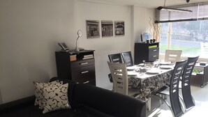 Un salon moderne avec un canapé noir, une table à manger dressée pour quatre personnes et un ordinateur portable posé sur la table. On y trouve également une bibliothèque remplie de divers objets et une fenêtre équipée de stores.