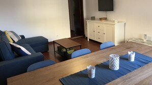 Een woonkamer met een bank, een houten salontafel, een eettafel met blauwe placemats, en een aan de muur bevestigde televisie.