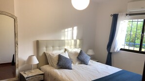 Een net opgemaakt bed met een wit hoofdbord en blauw beddengoed, een bedlampje en een nachtkastje met een lamp.