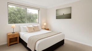 Une chambre avec une grande fenêtre, un lit recouvert de linge de lit blanc, deux tables de chevet avec des lampes et un tableau encadré au mur.