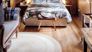 Quarto aconchegante com teto de madeira, uma cama grande com colcha estampada, duas mesas de cabeceira com abajures, um banco de madeira e uma cadeira.