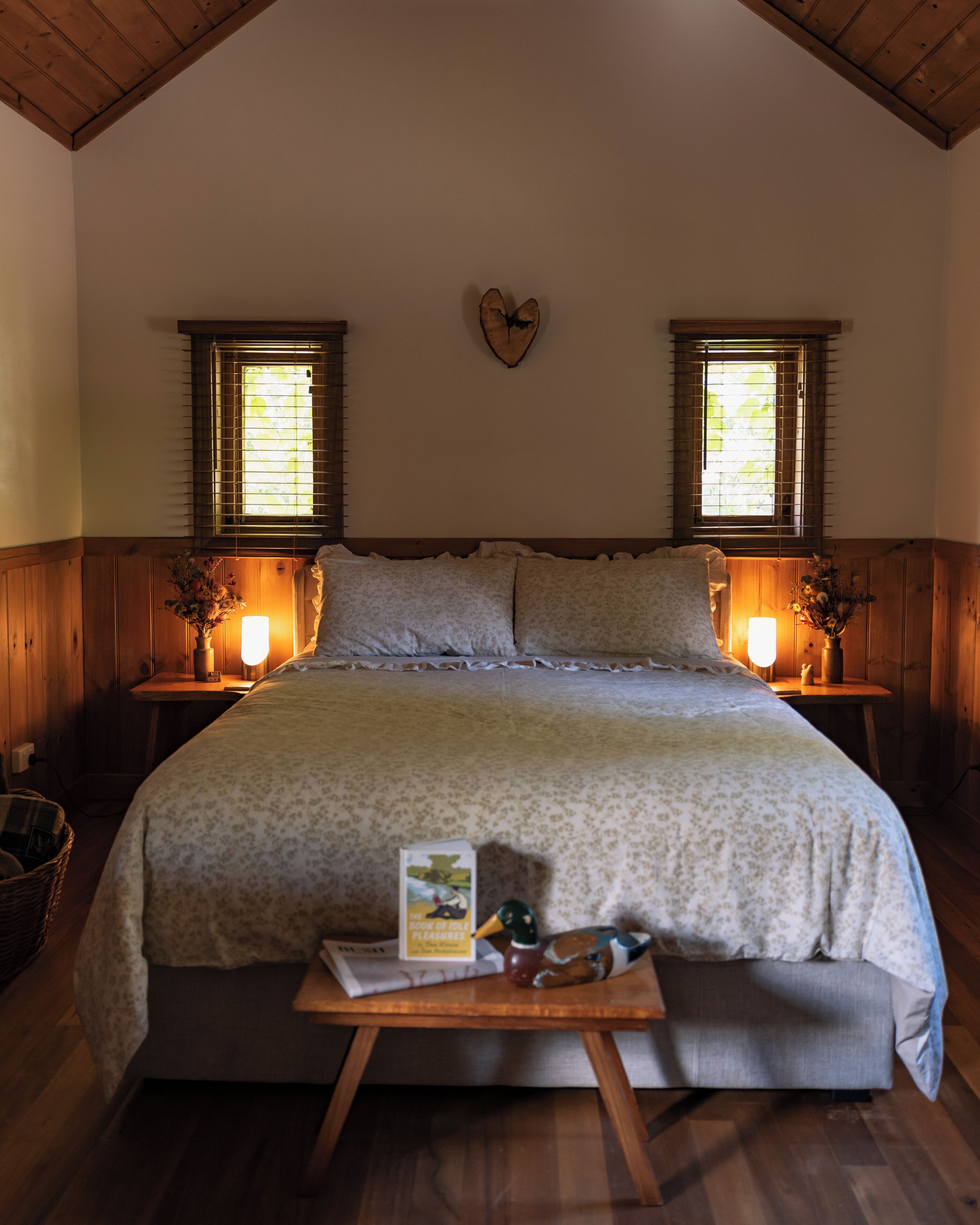 A bedroom with a large bed, wooden walls, two windows with blinds, bedside tables with lamps, and a heart-shaped wall decoration.