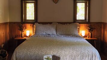 A bedroom with a large bed, wooden walls, two windows with blinds, bedside tables with lamps, and a heart-shaped wall decoration.