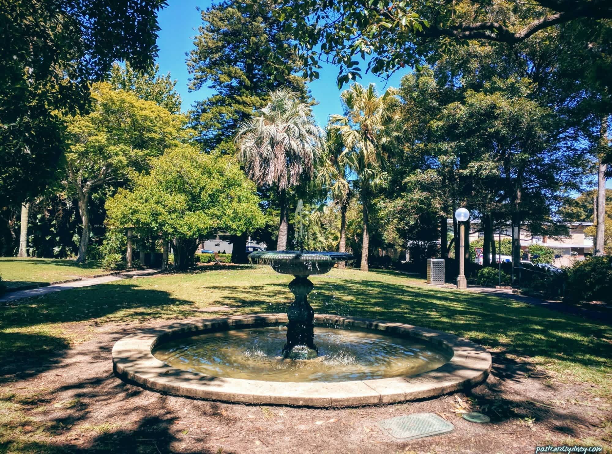 Ein kreisrunder Brunnen mit einer Fontäne in der Mitte, umgeben von einer Steinumrandung, eingebettet in einen Park mit Bäumen und blauem Himmel.