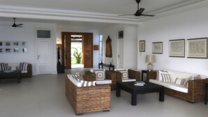 A spacious living room with a large sofa set, a coffee table, and a dining table. There are ceiling fans, framed pictures on the walls, and a door leading to a balcony.