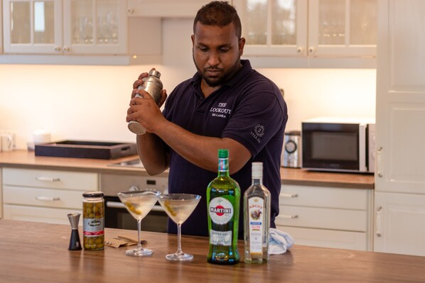 Una persona preparando una bebida en una cocina con armarios blancos, un microondas y varias botellas y vasos sobre la encimera.
