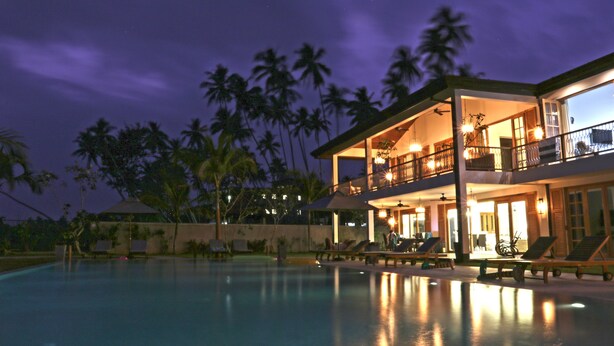 A two-story resort with a swimming pool, outdoor seating, and a view of palm trees at night.
