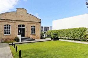 Um edifício com fachada de tijolos, uma estrutura branca moderna ao lado, e um gramado bem cuidado com um hedge.