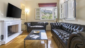 A living room with a fireplace, leather sofas, a flat-screen TV, and a view of a hill through open windows. - Aerides 3 Luxury Suites - the heart of the historic center, with Acropolis view (Athens)