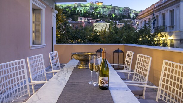 A terrace with a table set for two, a bottle of wine, and two glasses. The terrace overlooks a hillside with buildings and greenery. - Aerides 3 Luxury Suites - the heart of the historic center, with Acropolis view (Athens)