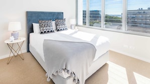 Ett modernt hotellrum med ett stort fönster, en säng med mönstrade kuddar, ett sängbord med en lampa och utsikt över stadsbilden.
