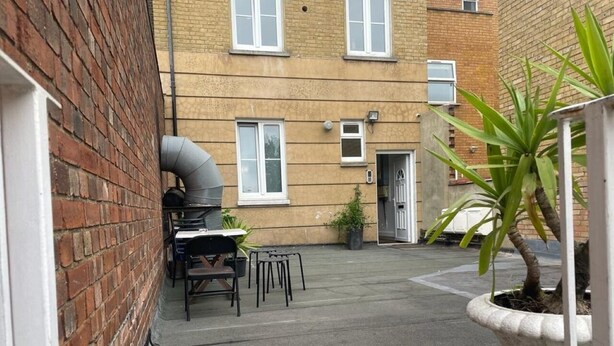 Une ruelle étroite, avec un mur de briques d’un côté et un bâtiment doté de fenêtres et d’une porte de l’autre. On y voit une petite table et des chaises disposées à l’extérieur, et un pot de fleurs est visible au premier plan.