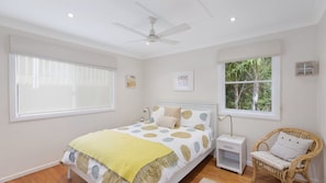Quarto com cama, mesa de cabeceira, cadeira, ventoinha no teto e duas janelas.