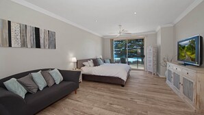 A bedroom with a bed, a dark-colored sofa, a television, and a view of a balcony with a pool. - Siesta by the Sea - Beachside Retreat (COPACABANA)