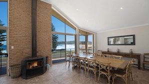 A spacious dining area with a large wooden table and chairs, a fireplace, and large windows offering a view of the outdoors. - Siesta by the Sea - Beachside Retreat (COPACABANA)