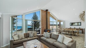 A spacious living room with a large window, a fireplace, and a dining area with wooden furniture. - Siesta by the Sea - Beachside Retreat (COPACABANA)