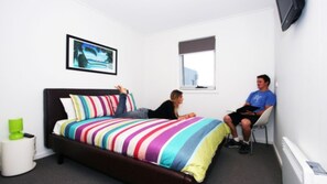 A bedroom with a bed, a window, a TV, and a green trash bin.