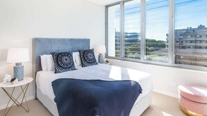Una habitación de hotel moderna con una cama grande, vistas a la ciudad y un otomán rosa redondeado.