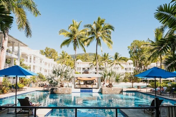 Zona de piscina con sillones reclinables, palmeras y un edificio al fondo.