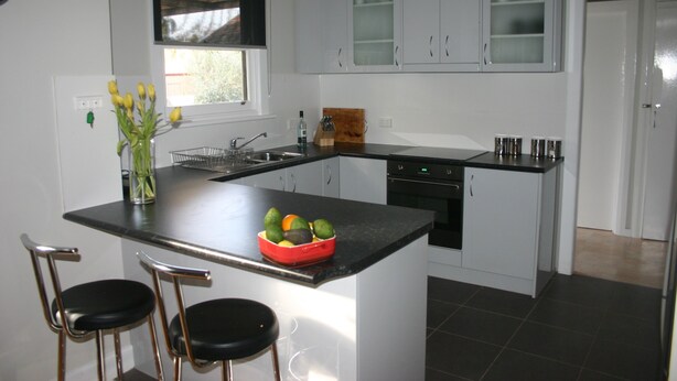 A modern kitchen with white cabinets, a black countertop, and a built-in oven. There are two bar stools and a vase with flowers on the counter. - Banksia Cottage - An Old Charm Cottage that has been completely renovated (Albury)