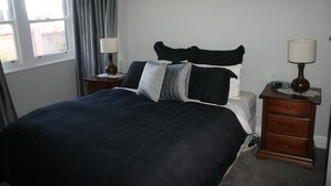 A neatly made bed with a dark bedspread and multiple pillows, a wooden nightstand with a lamp, and a window with curtains. - Banksia Cottage - An Old Charm Cottage that has been completely renovated (Albury)