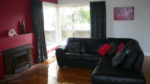 A living room with a black leather sofa, a fireplace, and a large window with curtains. - Banksia Cottage - An Old Charm Cottage that has been completely renovated (Albury)