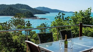 Una mesa con una botella y vasos en un balcón con vistas al agua y una colina boscosa.