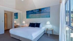 Habitación de hotel con una cama grande, una mesita de noche, una lámpara, un cuadro con una playa y una ventana con cortinas.