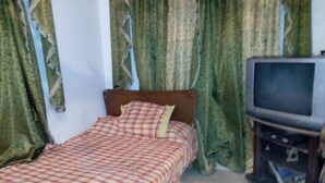 Ein kleines Zimmer mit einem Einzelbett, einem Fernseher auf einem Holzständer und grünen Vorhängen.
