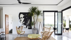 Un salón moderno con un cuadro abstracto grande, muebles de mimbre, una planta en maceta y vistas a través de un ventanal.