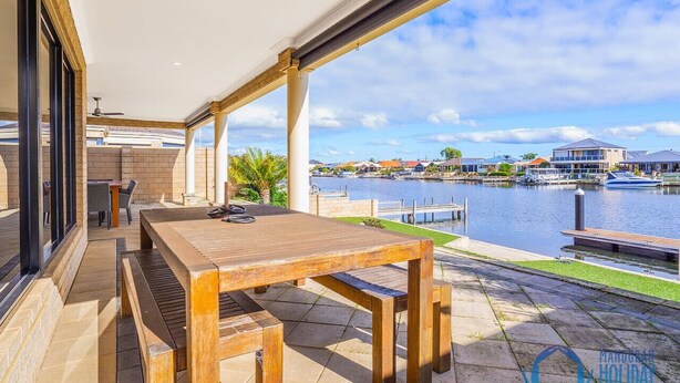 An outdoor patio with a wooden table and benches, overlooking a marina with boats and buildings in the background. - Canal Elegance in Yunderup (South Yunderup)