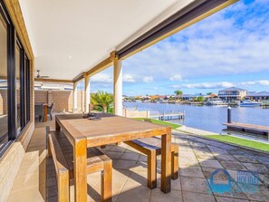 An outdoor patio with a wooden table and benches, overlooking a marina with boats and buildings in the background. - Canal Elegance in Yunderup (South Yunderup)