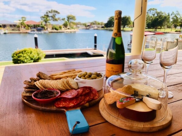 A wooden table with a variety of food items, including sliced meats, cheeses, and olives, set against a scenic lakeside view. - Canal Elegance in Yunderup (South Yunderup)