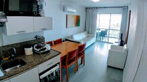 A compact kitchen with a microwave, coffee maker, and dishwasher. There is a dining area with a table and chairs, and a white sofa. A large window provides a view of the outdoors. - VistaMar at Villa del Sol Residences (Recreio dos Bandeirantes)