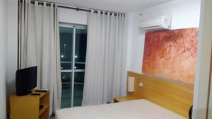 A hotel room with a bed, a TV on a wooden stand, a window with curtains, and an air conditioning unit on the wall. - VistaMar at Villa del Sol Residences (Recreio dos Bandeirantes)