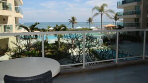 A balcony with a view of a pool and the ocean, surrounded by palm trees and umbrellas. - VistaMar at Villa del Sol Residences (Recreio dos Bandeirantes)