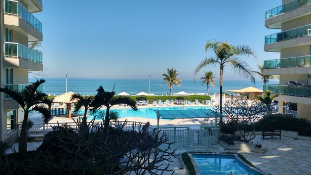 A resort pool area with a view of the ocean, palm trees, and a clear sky. - VistaMar at Villa del Sol Residences (Recreio dos Bandeirantes)